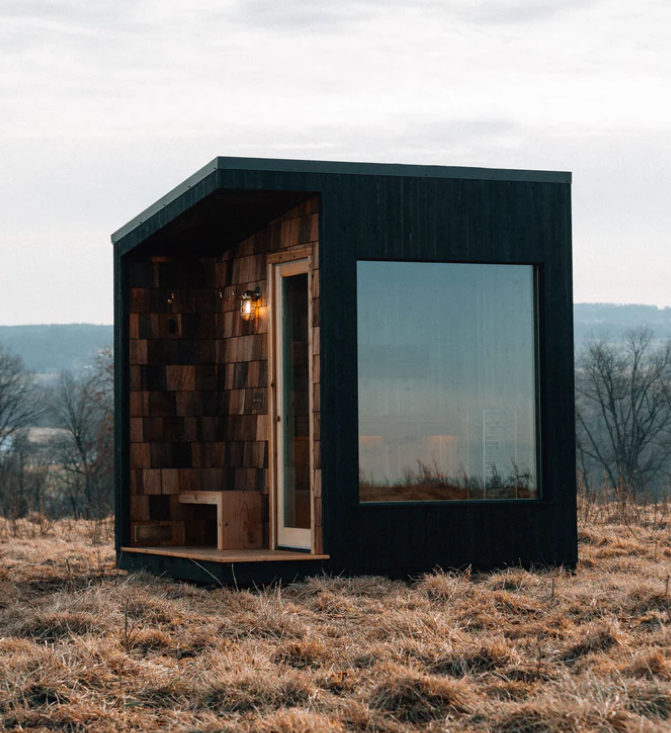 Dark modern shed-roof sauna with large glass window in a dry field at dusk